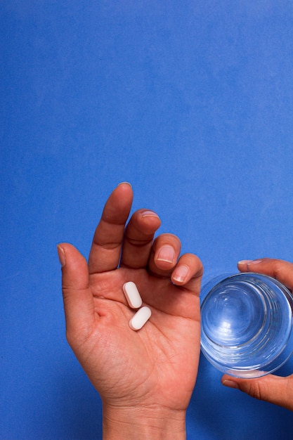 Hand holding white pills on blue background and a glass of water. Copy space.