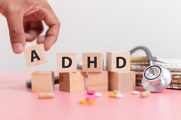 Photo hand holding wood cube block abbreviation of adhd with stethoscope and pills on pink table attention deficit hyperactivity disorder adhd concept