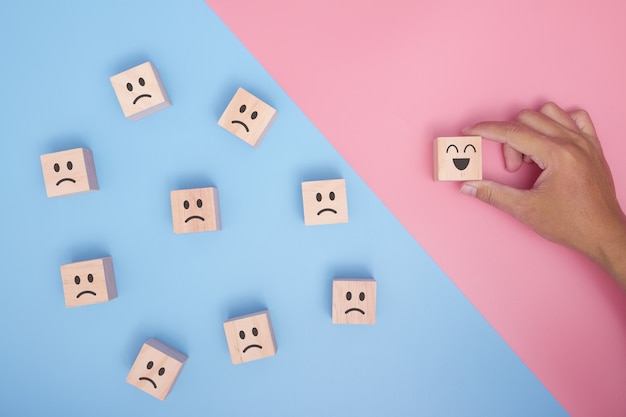 Hand holding wood cube with smiley face icon symbol on blue and pink background. Customer service rating, Satisfaction survey concept.