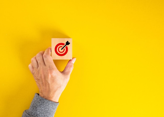 Hand holding wooden block with dartboard with arrow on yellow background