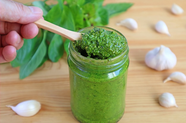 Mano che tiene il cucchiaio di legno che raccoglie il pesto di basilico fresco o il pesto alla genovese dalla bottiglia