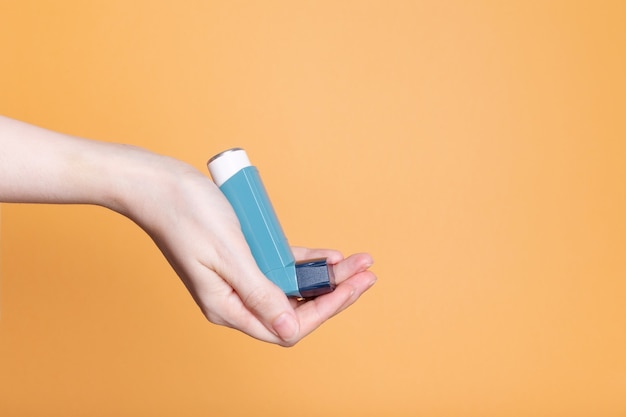 Hand holds blue inhaler to treat asthma on orange background