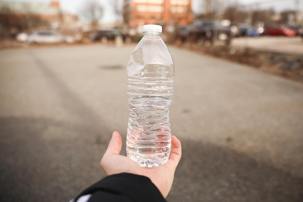 A hand holds a bottle of water in front of a parking lot.