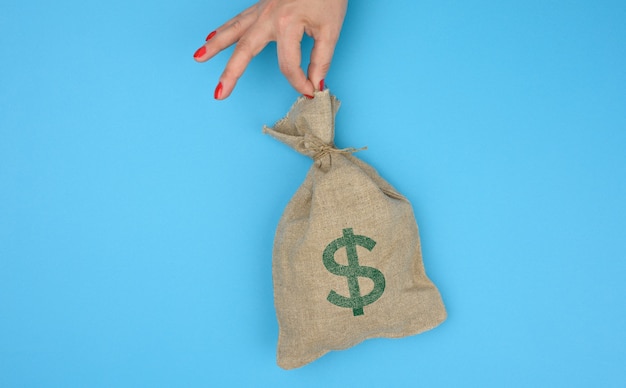 Photo hand holds a canvas bag full of money with a us dollar icon on a blue background. budget planning