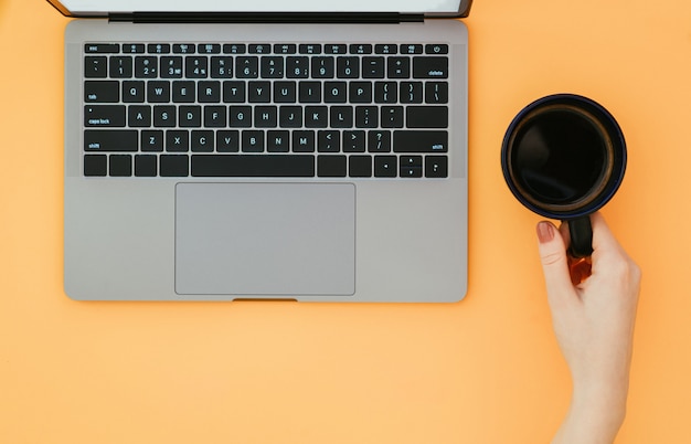 Hand holds a cup of coffee and a laptop on an orange surface