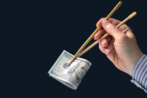 Hand holds dollars with bamboo chopsticks on a dark background financial industry in food cash spending concept