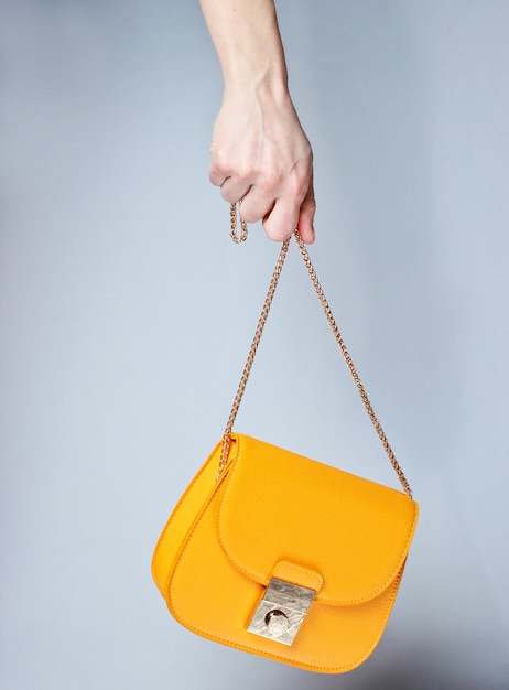 Hand holds fashionable yellow leather bag with golden chain on gray surface