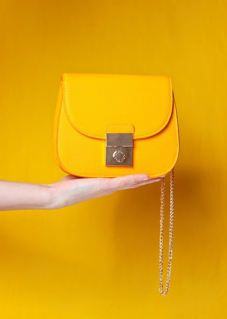 Hand holds fashionable yellow leather bag with golden chain on yellow surface