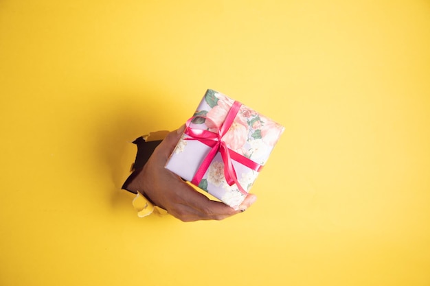 A hand holds a gift box a torn yellow paper wall