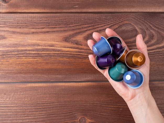 A hand holds a handful of colorful coffee capsules over a brown wooden background. Top view, copy space,