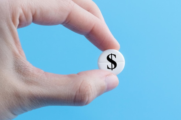 A hand holds a medical pill with a dollar currency sign on a blue background The concept of a rise in the price of medicines