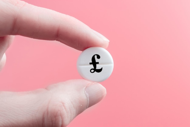 A hand holds a medical pill with an English pound sterling currency sign on a pink background The concept of a rise in the price of medicines in Great Britain