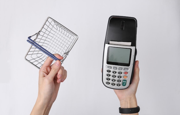 Hand holds payment terminal and supermarket basket on white background Payment for purchases