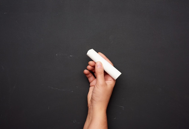 Hand holds a piece of white chalk on the background of an empty black chalk board