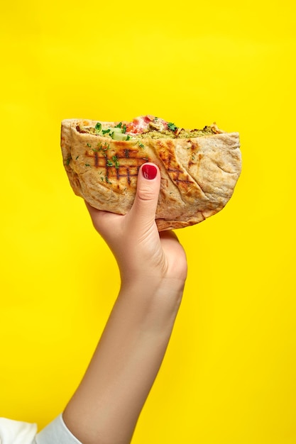 Hand holds pita with chicken and vegetables on a yellow background. Shawarma with chicken