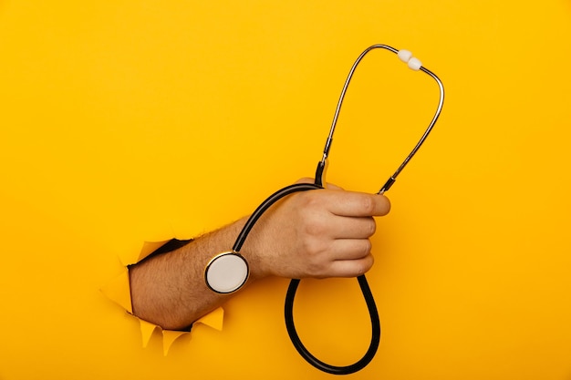 Hand holds a stethoscope out of a hole torn in yellow paper wall with copy space