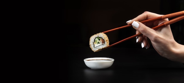 Hand holds sushi over bowl with soy sauce on black table.