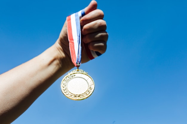 A hand holds up a first-place gold medal, with the sky in the background. Victory concept