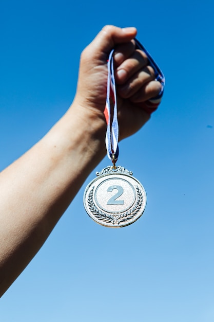 A hand holds up a second-place silver medal, with the sky in the background. Victory concept