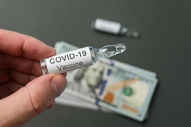 Hand holds a vial with label Covid-19 on money background. Cost of vaccine for 2019-nCov infectious disease.