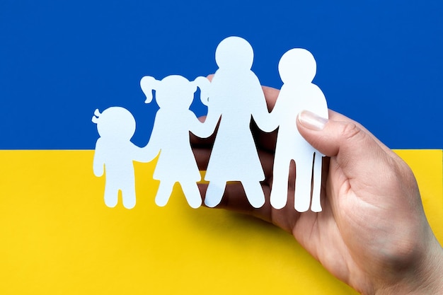 Hand holds white paper silhouette of Ukrainian family Flat lay top view on blue yellow paper background Ukraine flag colors Volunteer help to refugees from Ukraine