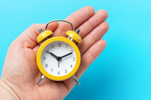 Hand holds yellow alarm clock on a blue surface, copy of space