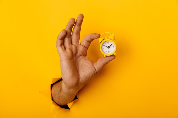 Photo hand holds yellow alarm clock through a paper hole in yellow background