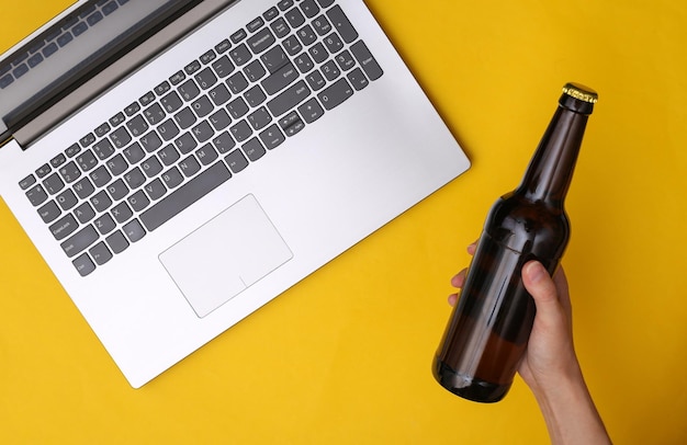 Hand houdt een flesje bier op een gele achtergrond met een laptop. Het concept van rust, ontspanning na een dag hard werken. Bovenaanzicht