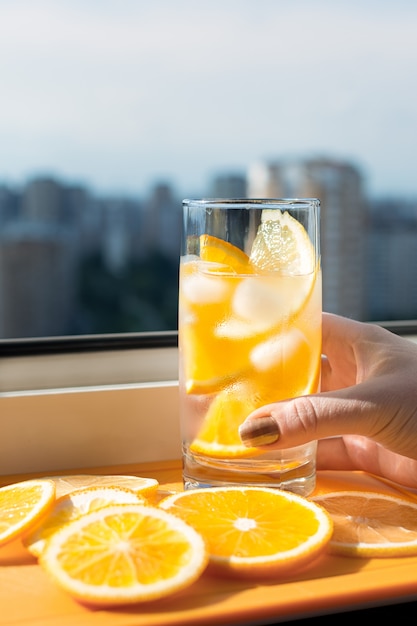 Hand houdt een glas met een drankje met ijs en stukjes sinaasappel op de achtergrond van de stad