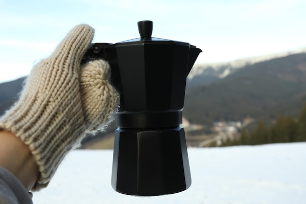 Фото Рука в рукавице держит кофеварку против гор