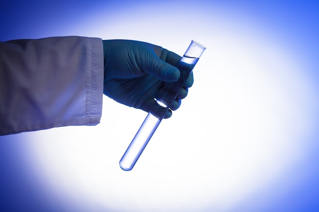 Фото Рука в резиновой перчатке держит пробирку с прозрачной жидкостью на синем фоне