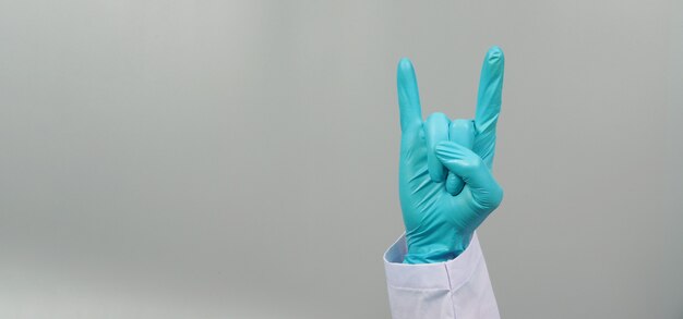 La mano è fare il segno della mano con le corna del diavolo e indossare un camice da medico e un guanto medico blu su sfondo grigio. riprese in studio.