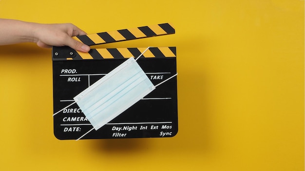 The hand is holding a black clapper board with a medical face mask on yellow background.