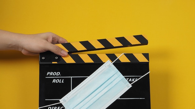 Photo hand is holding black and yellow color clapper board or movie slate with face mask. it use in video production and cinema industry on yellow background.