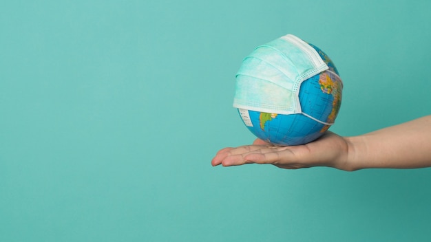 Foto la mano tiene il globo terrestre e la maschera facciale su sfondo verde o tiffany blue. concetto covid-19.