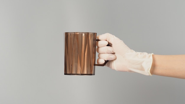 Hand is holding plastic cup and wear latex glove on grey background.