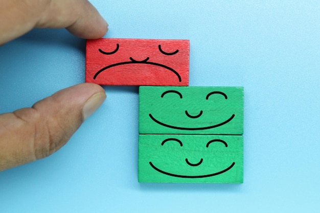 A hand is holding a red and green wooden smiley face between two green blocks.