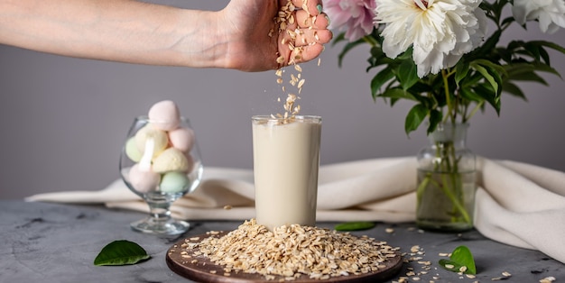 A hand is spilling oat flakes into a glass of natural oat milk on the table