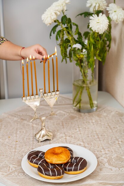 ユダヤ人の女性の手が、皿のドーナツの隣のテーブルにあるハヌカのろうそくに火を灯します