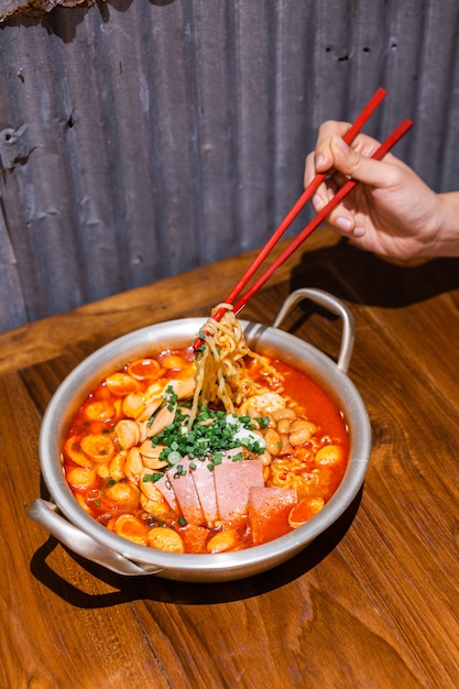 Hand knijpen noedels met stokjes. Traditionele Koreaanse ramen soep met kimchi, ham, worst en kaas in zilveren kom op houten tafel. Koreaanse keuken. Lekker Aziatisch eten.