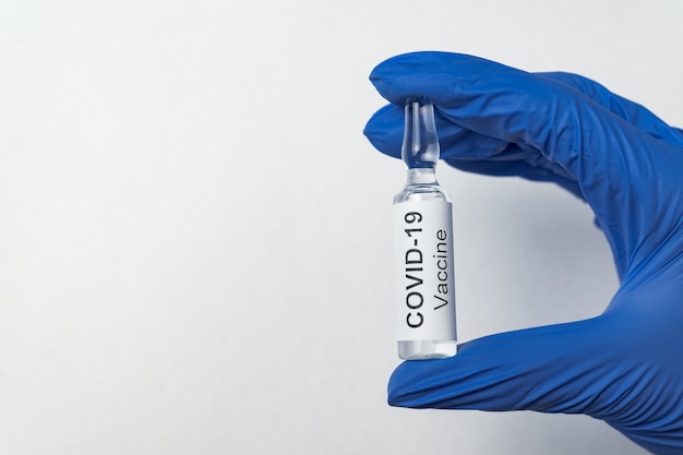 Hand in latex glove holding vial with label Covid-19. Ampoule with cure for coronavirus, on white background.