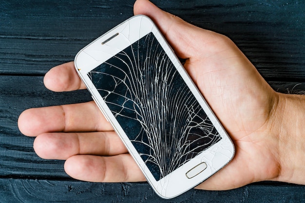 Hand of a male holding white smartphone with damaged glass\
screen indoors broken sensory gadget in the man\'s hand on the black\
wooden background closeup