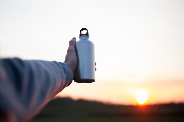 Hand of man holding aluminium bottle for water, on background of sunset