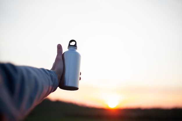 Mano dell'uomo che tiene la bottiglia di alluminio per l'acqua, sullo sfondo del tramonto