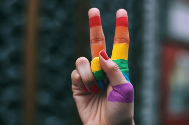 The hand of a man making the V sign with his fingers painted as the rainbow flag against a blue background with some blank space on top