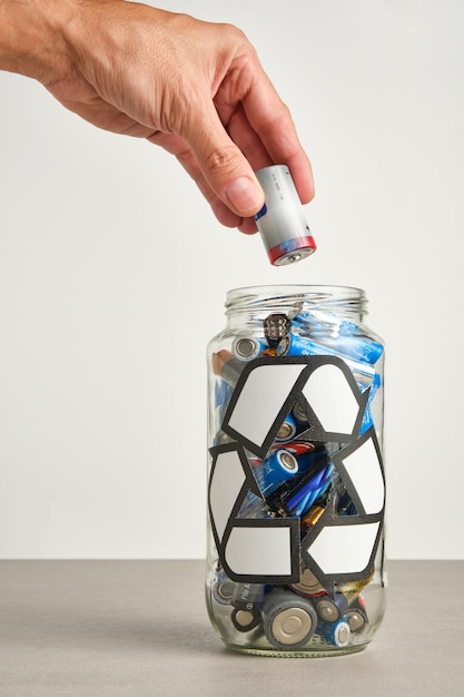 Hand of a man pouring a used battery into a glass jar with the recycling symbol full of used batteries