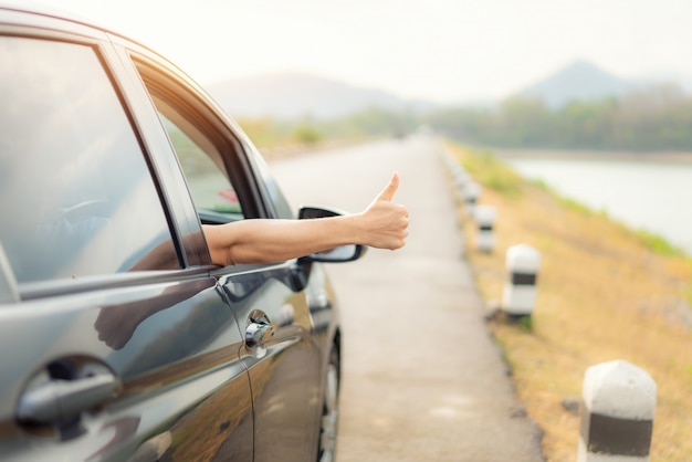 La mano del viaggiatore dell'uomo con i pollici su dall'automobile forma la strada asfaltata va viaggiare con il lago e le montagne del bello paesaggio e la luce solare nella sera.