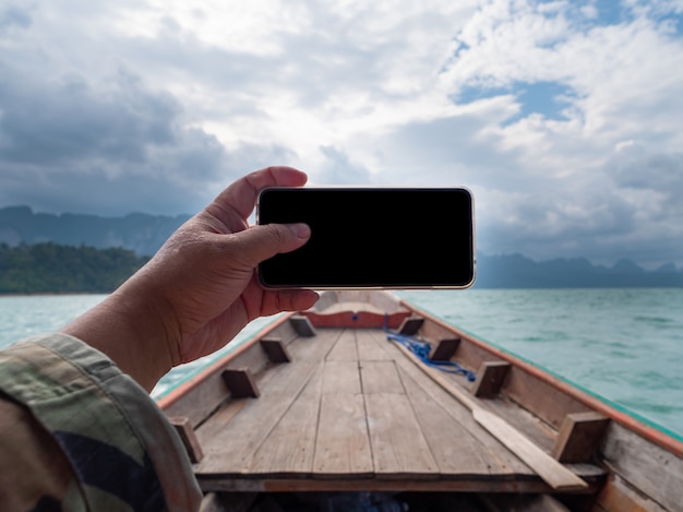 Hand of man using mobile smart phone against scenic landscape of boat view in the big river and reservoir dam with mountain and nature forest