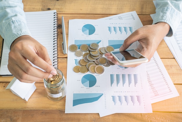 Hand of man using smartphone and putting coin into the glass on wooden working table with a pile of coins, chart notebook, pen and mini model white house - Investment, Business, Finance and Banking