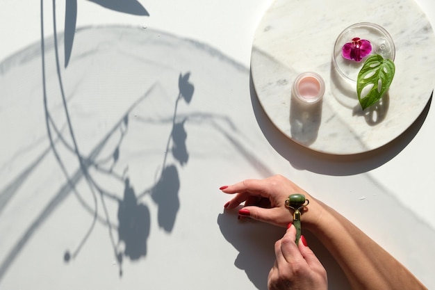 Photo hand massage, lymph drainage with green jade stone roller. moisturizing cream on marble plate. exotic flat lay with monstera leaves, magenta orchid flowers. sunlight, long shadows.